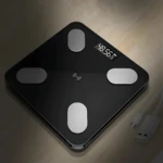 Báscula de grasa corporal negra con pantalla digital mostrando 48.56 kg, cuatro sensores metálicos y cable USB incluido, vista cenital sobre superficie de madera.