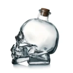 Botella de vidrio transparente con forma de calavera, tapada con corcho, ideal para servir bebidas en eventos temáticos.