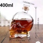 Botella de vidrio con forma de calavera, capacidad de 400 ml, mide 13.5 cm de alto y 11.4 cm de ancho, contiene líquido marrón, ideal para servir bebidas en fiestas.