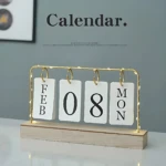 Calendario de escritorio con base de madera clara y marco dorado con luces LED. Muestra fichas blancas indicando la fecha 08 de febrero y el día lunes. Diseño minimalista para decoración.