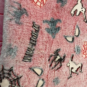 Detalle de cobija de felpa rosa con estampado de Spiderman. Se observa textura suave y diseños de araña, máscara y telaraña en relieve sobre fondo melocotón.