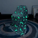 Cobija de felpa gris oscuro con estampado de lunas y estrellas brillando en verde neón, colgada sobre una silla en ambiente oscuro para demostrar su brillo.