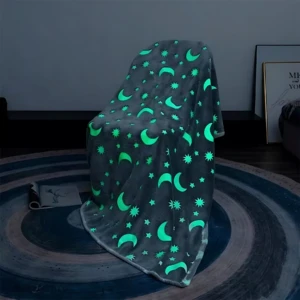 Cobija de felpa gris oscuro con estampado de lunas y estrellas brillando en verde neón, colgada sobre una silla en ambiente oscuro para demostrar su brillo.
