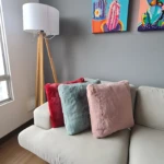 Cojín decorativo peluche largo suave en tonos rosa, azul y rojo sobre sofá blanco, con lámpara de pie y cuadros al fondo.
