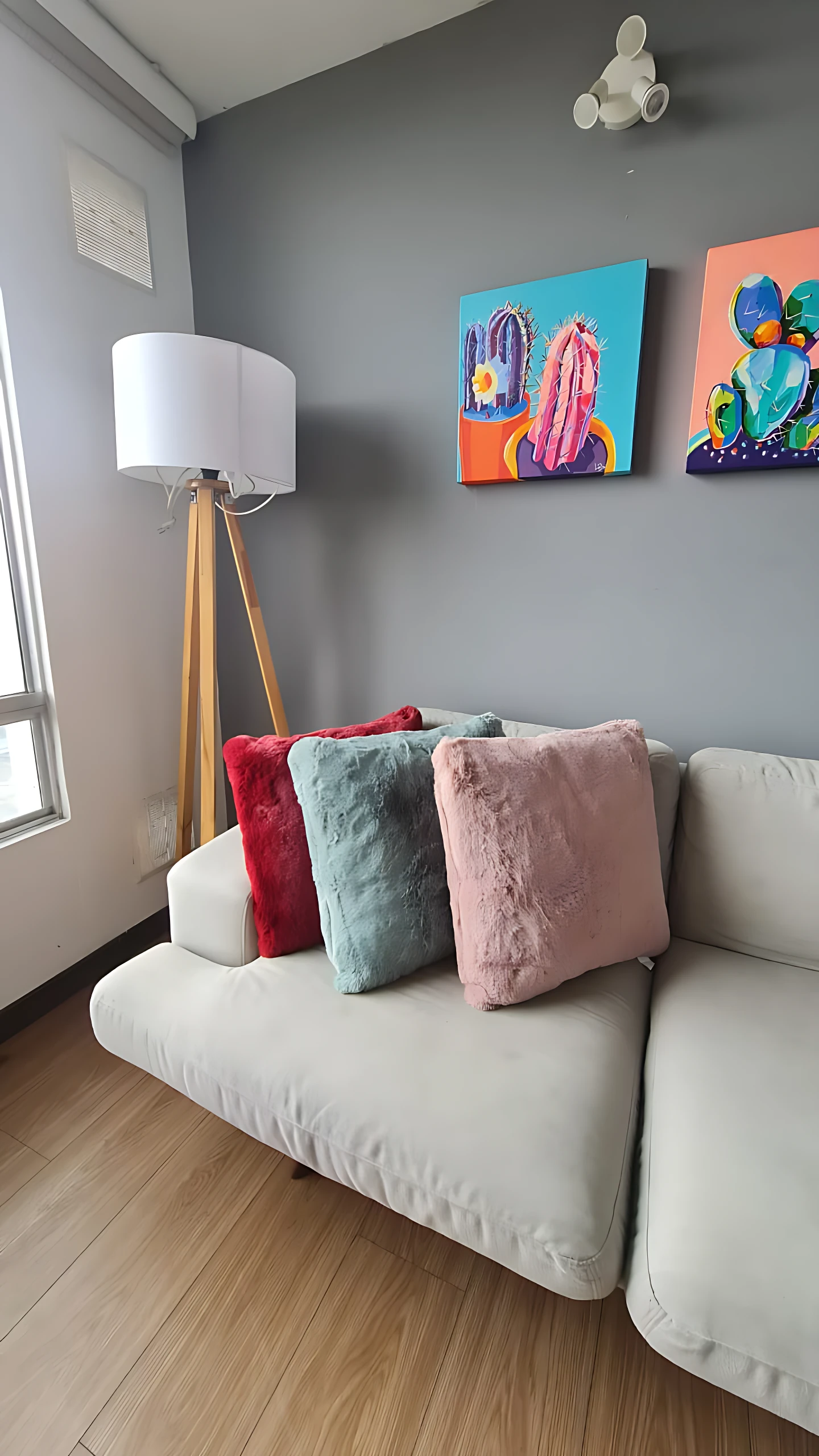 Cojín decorativo peluche largo suave en tonos rosa, azul y rojo sobre sofá blanco, con lámpara de pie y cuadros al fondo.