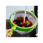 Colador giratorio verde con mango plegable, en uso bajo chorro de agua, lavando fresas y moras en colador blanco con agujeros redondos.