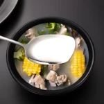 Cuchara de acero inoxidable con sopa de maíz y carne, servida en un tazón negro.