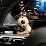 Peluche perro con gafas de piloto, color beige y marrón, colocado en el tablero de un vehículo.
