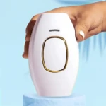 Depiladora láser IPL blanca con borde dorado, sostenida por mano con uñas blancas, sobre pedestal azul.