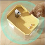 Dispensador automático de agua amarillo con grilla metálica, en uso por un gato naranja sobre piso de madera.