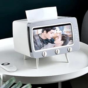 Dispensador de servilletas blanco y gris con pantalla de TV antigua mostrando pareja, soporte para móvil y control remoto.