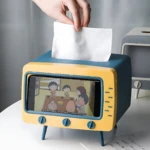 Dispensador de servilletas amarillo y azul con pantalla de TV antigua, sostiene móvil y servilletas.