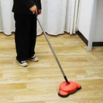 Escoba barredora manual naranja, con mango metálico, ideal para limpiar pisos de madera y rincones.
