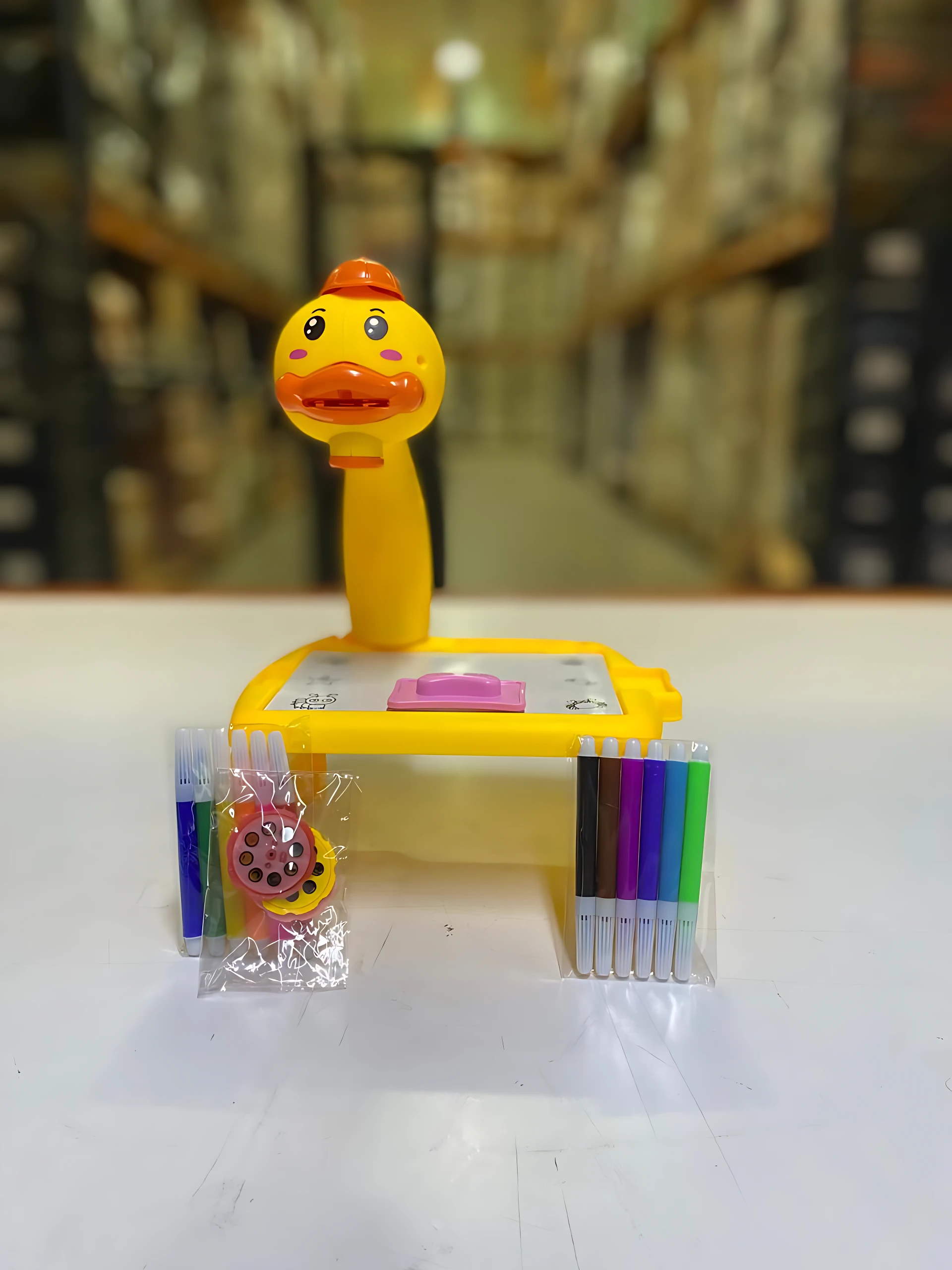 Escritorio amarillo con figura de pato naranja, incluye set de marcadores de colores y estuche con accesorios, sobre superficie blanca.