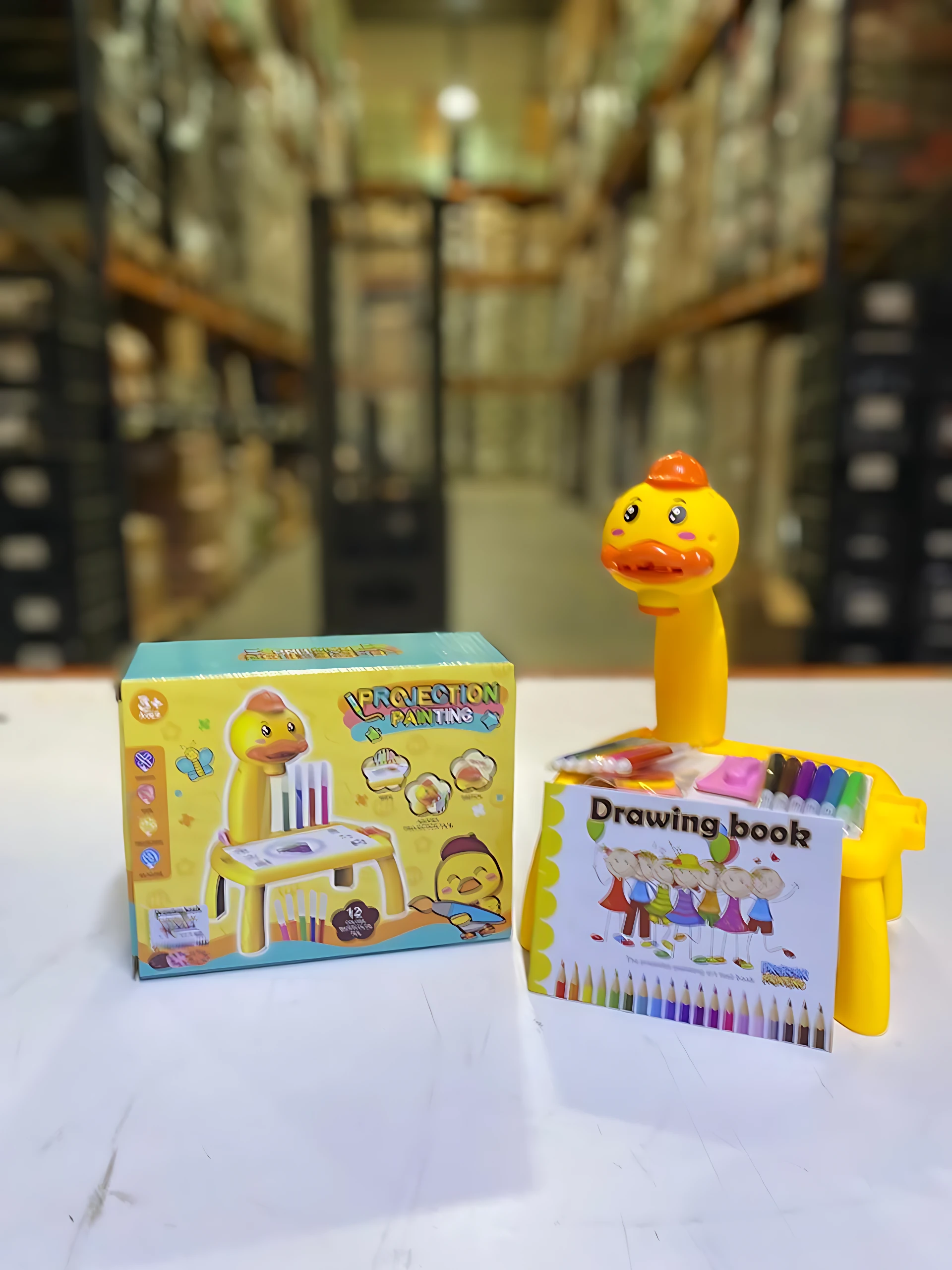 Escritorio amarillo con pato parlante, libro de dibujo y lápices de colores incluidos, sobre superficie blanca en almacén.