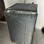 Forro protector gris para lavadora frontal, con cierre superior y paneles laterales acolchados, instalado sobre electrodoméstico en ambiente doméstico.
