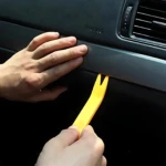 Herramienta plástica amarilla extrayendo moldura de tablero de auto, con mano aplicando presión y otra sosteniendo el mango.