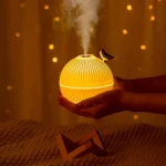 Humidificador difusor aroma pájaro color amarillo, con luz cálida y vapor saliendo de su parte superior, sostenido por manos sobre cama con cojines.