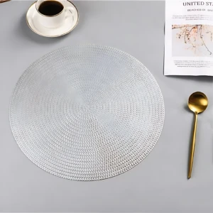 Placemat redondo tejido en color blanco, con textura de trenzado visible, acompañado de taza de café, cuchara dorada y revista sobre superficie gris clara.