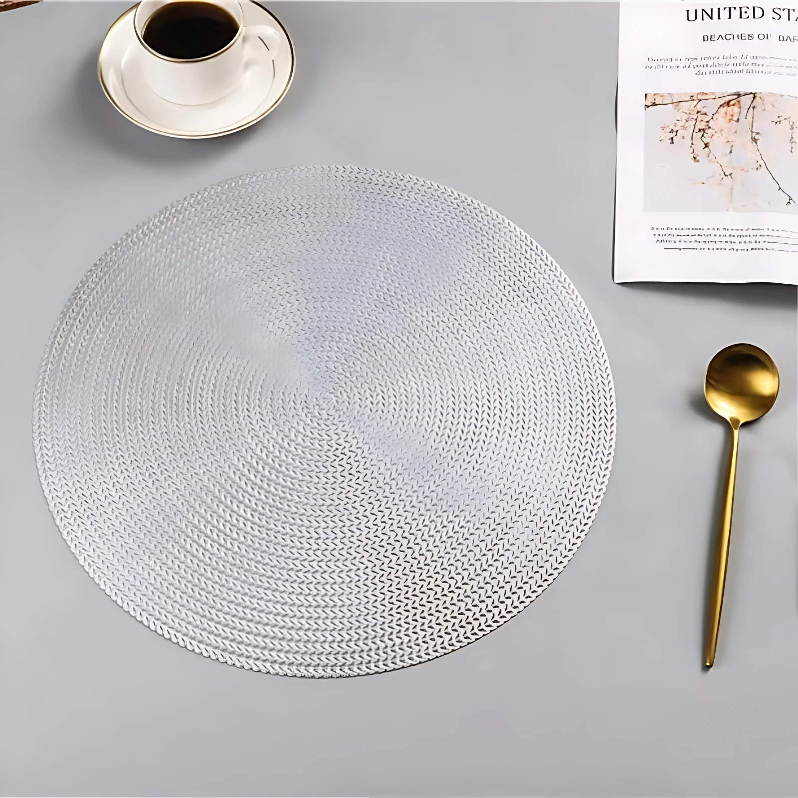 Placemat redondo tejido en color blanco, con textura de trenzado visible, acompañado de taza de café, cuchara dorada y revista sobre superficie gris clara.