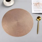 Placemate redondo tejido en tono rosa dorado, con textura de trenzado visible, acompañado de cuchara dorada y taza de café en esquina superior.