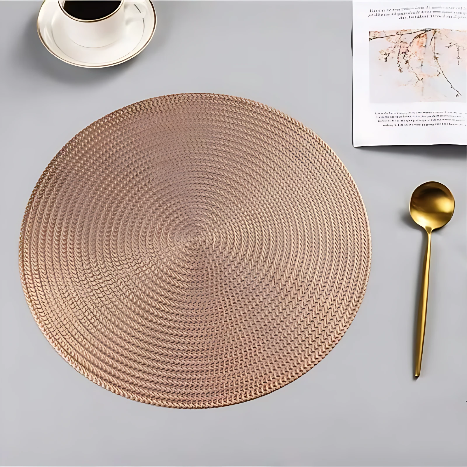 Placemate redondo tejido en tono rosa dorado, con textura de trenzado visible, acompañado de cuchara dorada y taza de café en esquina superior.