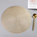 Placemate redondo tejido en color beige, con textura de trenzado visible. Acompañado de cuchara dorada y papel impreso en esquina superior derecha.