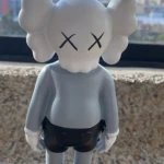 Figura decorativa de resina estilo KAWS, color gris claro con detalles negros, ojos en forma de X y zapatos brillantes, vista frontal sobre fondo urbano.