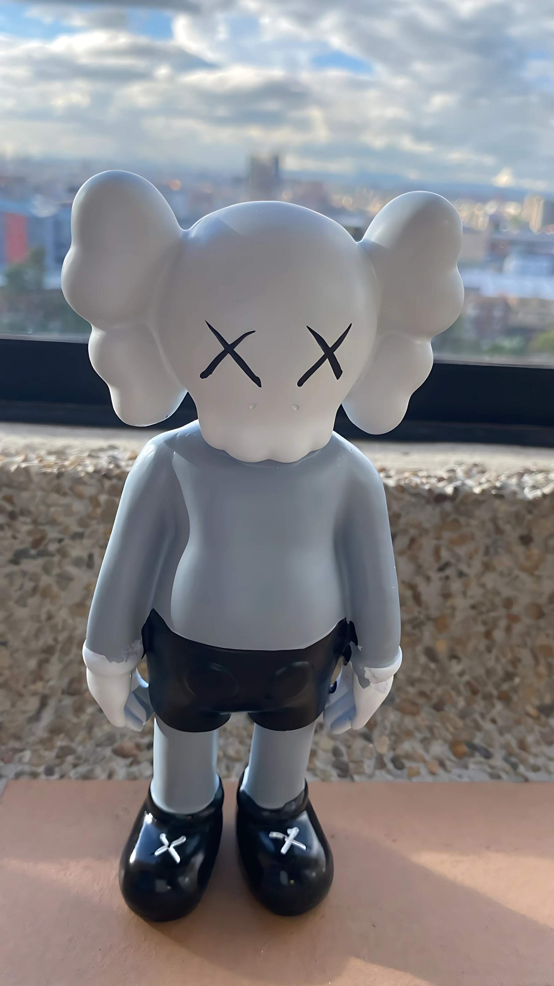 Figura decorativa de resina estilo KAWS, color gris claro con detalles negros, ojos en forma de X y zapatos brillantes, vista frontal sobre fondo urbano.