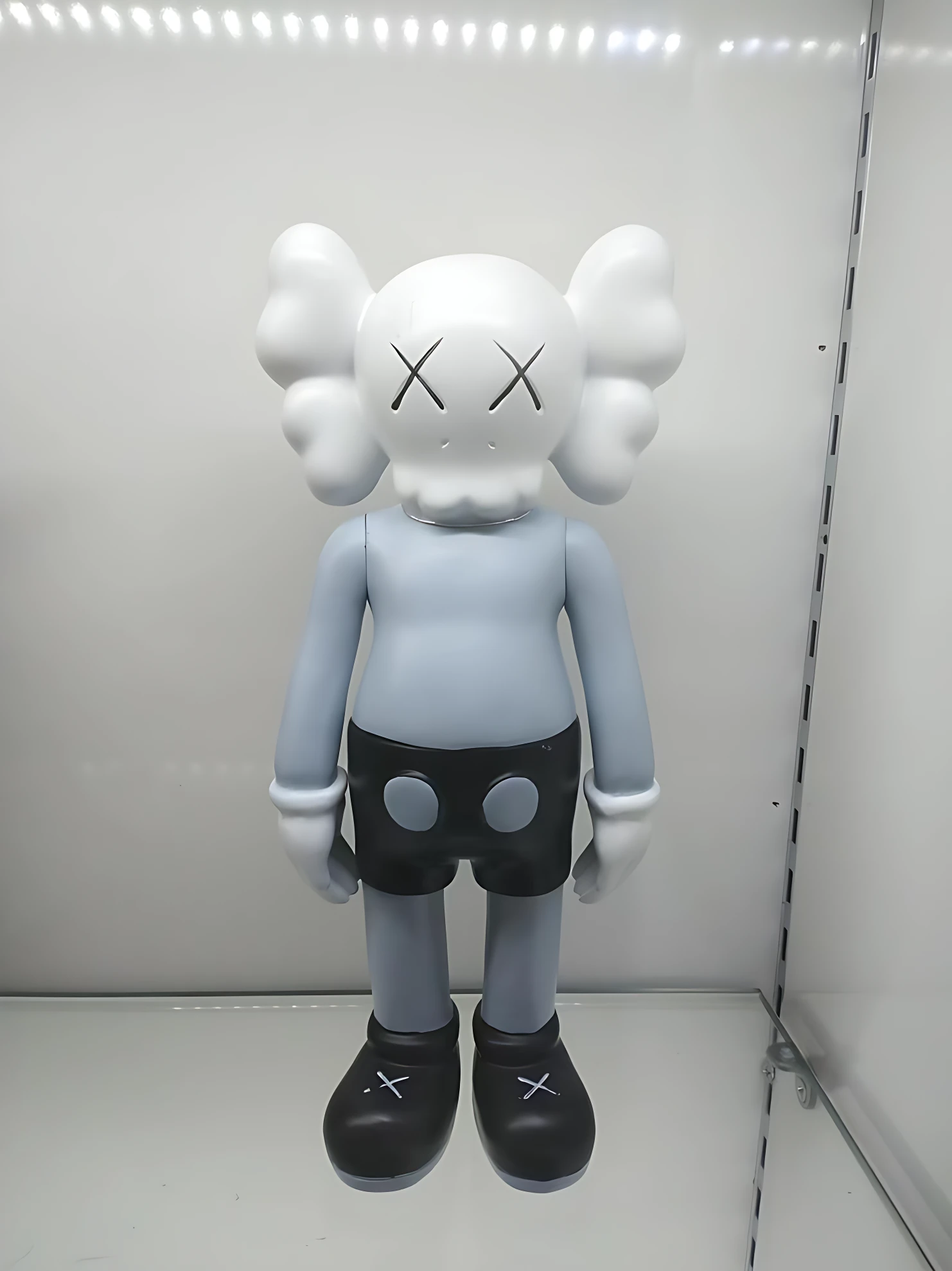 Figura decorativa de resina estilo KAWS, 18 cm, color gris claro con detalles negros y ojos en X, sobre fondo blanco.