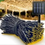 Cable de luces LED solares exteriores con 100 bombillos amarillos, panel solar negro y conectores negros, enrollado sobre fondo blanco con jardín iluminado al fondo.