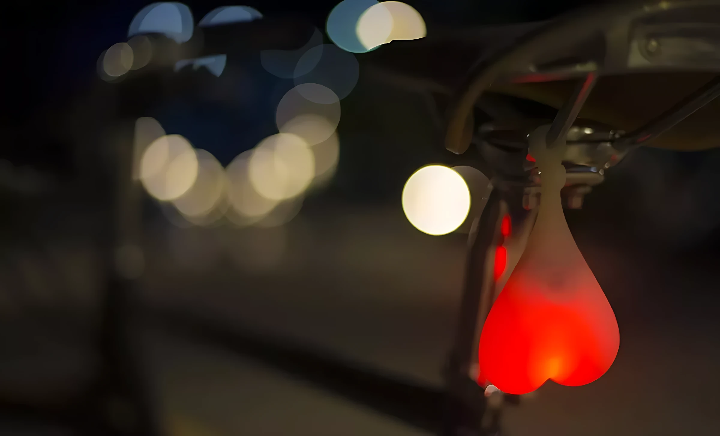 Set x2 luces LED rojas en forma de corazón para bicicleta, colgadas bajo el sillín, con fondo de luces nocturnas difuminadas.