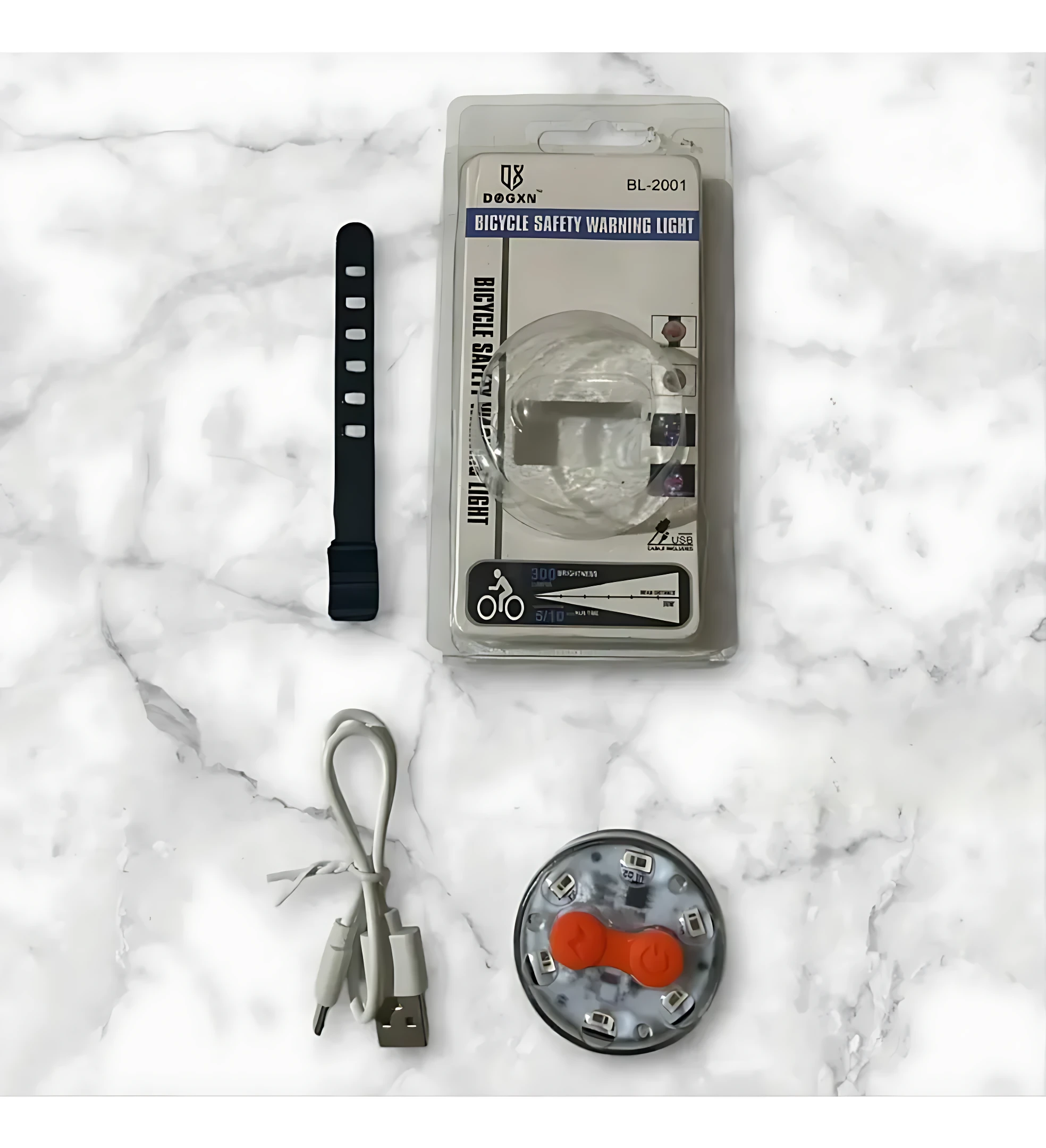 Pack de luces traseras para bicicleta recargables por USB, modelo BL-2001. Incluye luz circular con dos botones naranjas, correa negra con velcro y cable de carga. Presentación en blister transparente sobre fondo de mármol blanco.