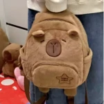 Mochila escolar infantil de felpa color marrón claro con diseño de capibara kawaii, mostrando rostro frontal y estuche frontal con logo.