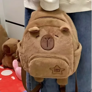 Mochila escolar infantil de felpa color marrón claro con diseño de capibara kawaii, mostrando rostro frontal y estuche frontal con logo.