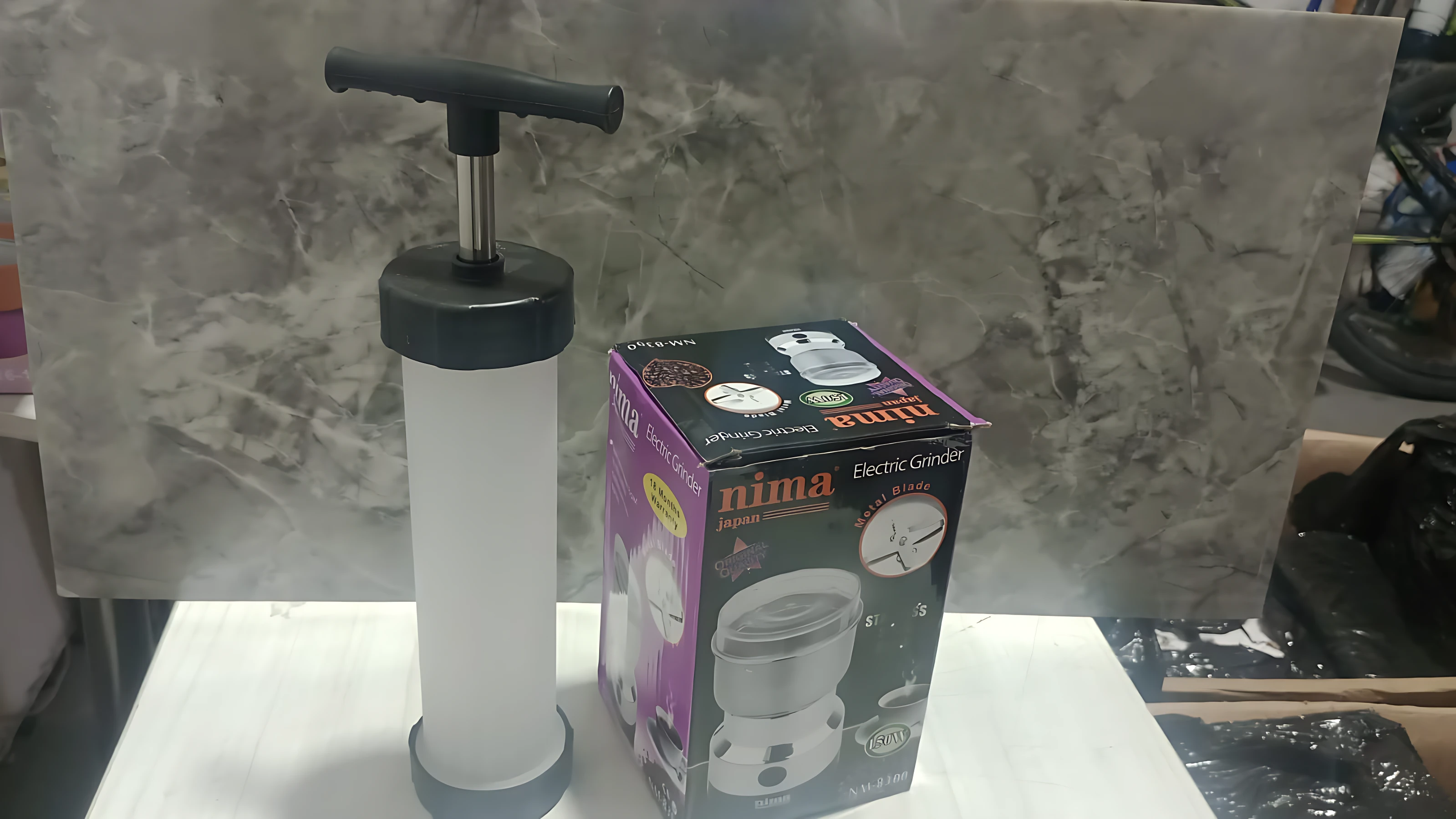 Molino de café y embutidora de salchicha en caja morada con del producto, junto al accesorio de bombeo manual negro y blanco sobre superficie blanca.