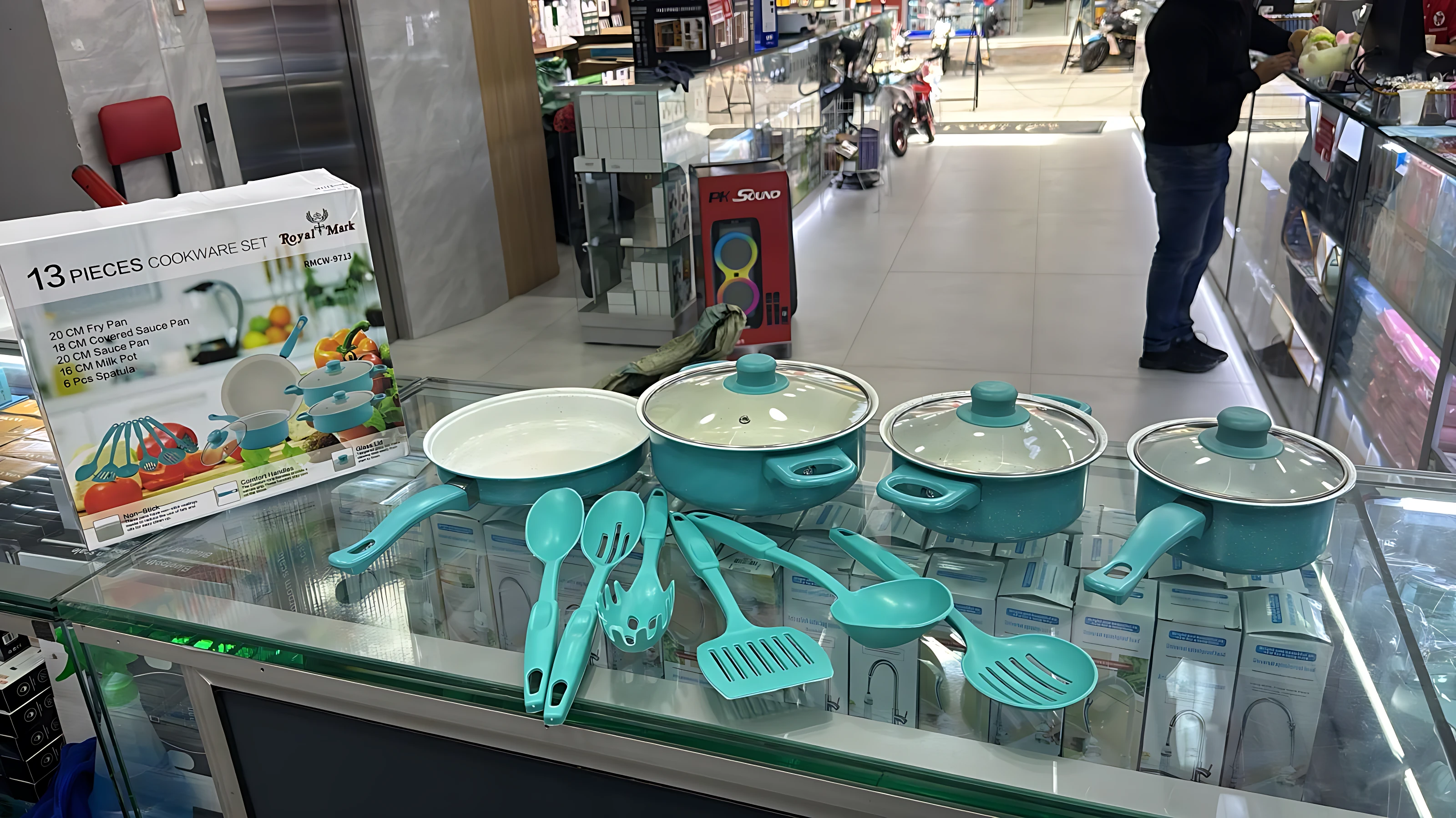 Set de cocina en color turquesa con ollas, sartén y utensilios sobre mostrador de vidrio en tienda. Caja visible al lado.