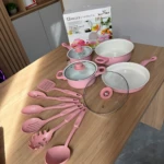 Set de cocina rosa con ollas, sartenes y utensilios de silicona sobre mesa de madera, junto a su caja original de 13 piezas.