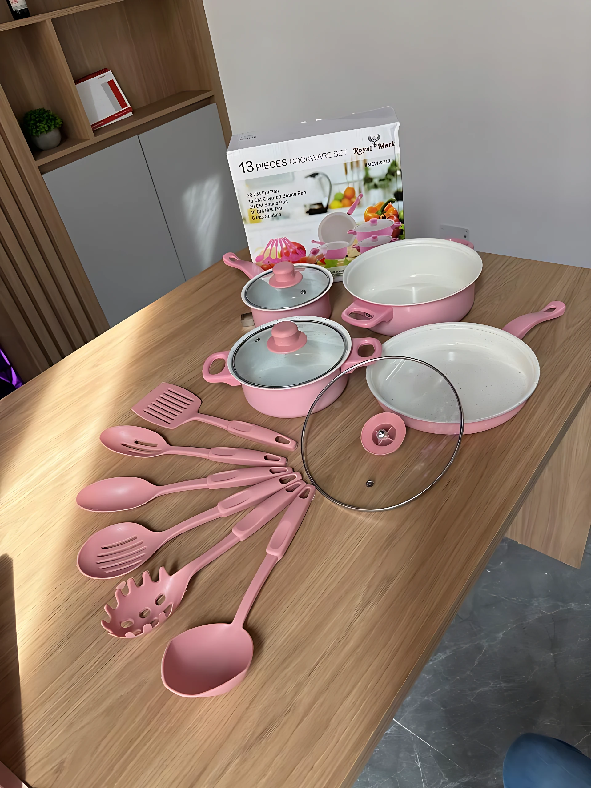 Set de cocina rosa con ollas, sartenes y utensilios de silicona sobre mesa de madera, junto a su caja original de 13 piezas.