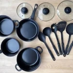 Set de sartenes negras con tapas de vidrio y utensilios de cocina en tonos oscuros, dispuestos sobre superficie de madera clara.