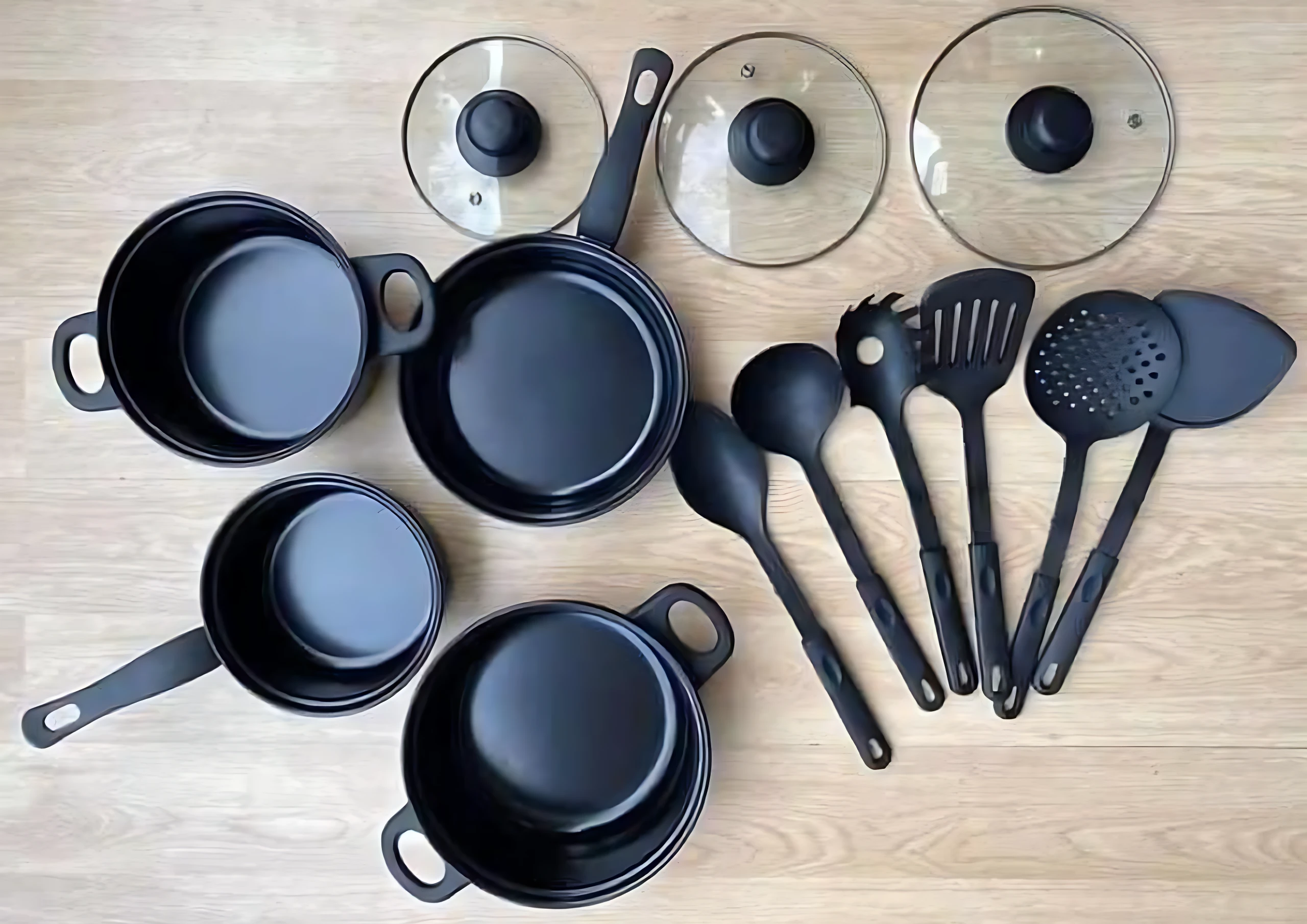 Set de sartenes negras con tapas de vidrio y utensilios de cocina en tonos oscuros, dispuestos sobre superficie de madera clara.