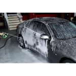 Pistola para mangueras 4 en 1 con jabón integrado en uso, aplicando espuma blanca sobre carrocería de auto gris en lavado manual.