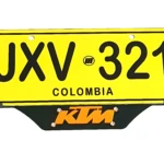 Portaplaca de acero inoxidable KTM en color negro con logo naranja, diseñado para montar placa amarilla de Colombia JXV 321.