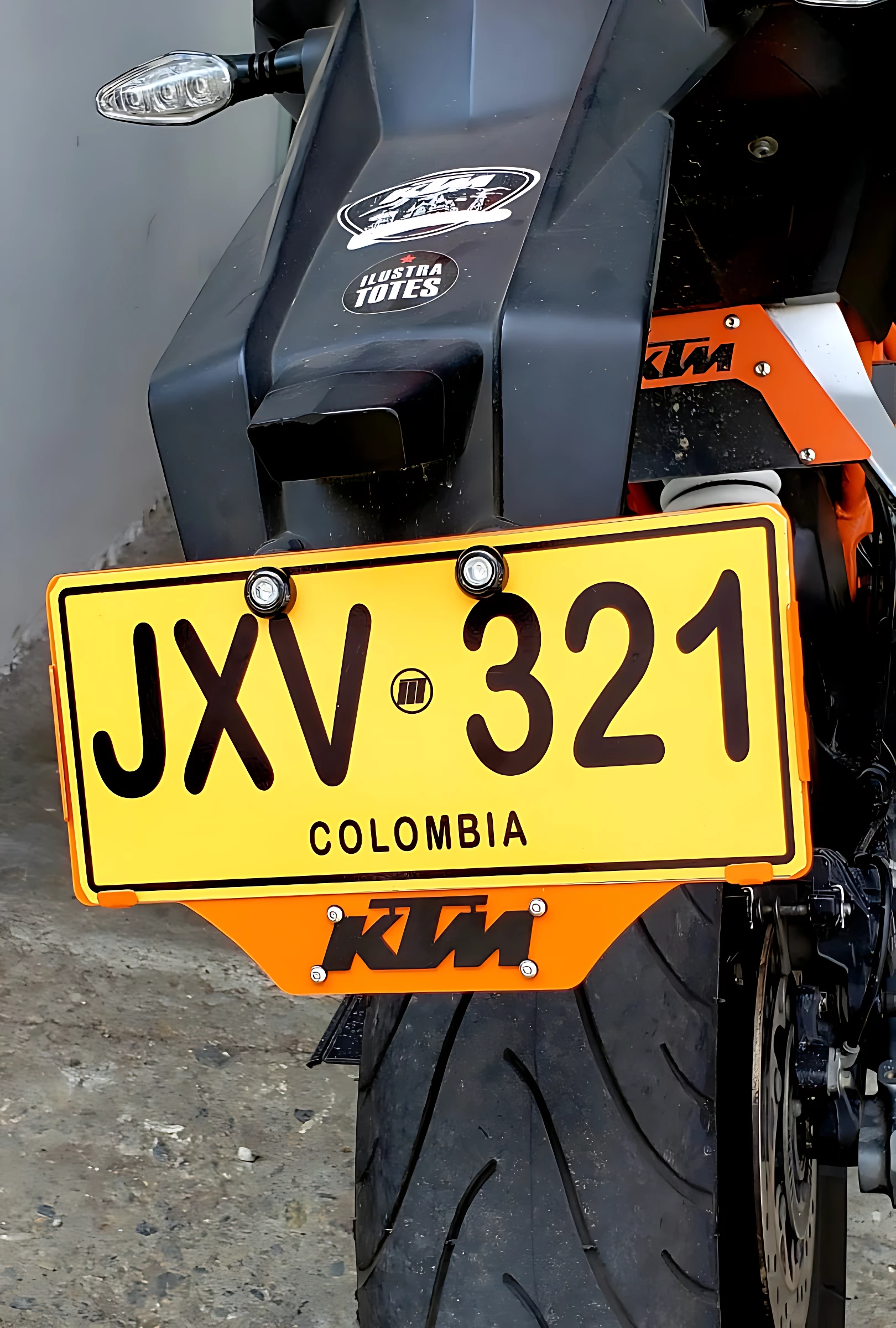Portaplaca KTM 3D en acero inoxidable, color naranja brillante, montado en la parte trasera de una motocicleta. Placa amarilla con texto negro JXV 321 y leyenda COLOMBIA. Incluye logo KTM en la base y detalles de fijación visibles.