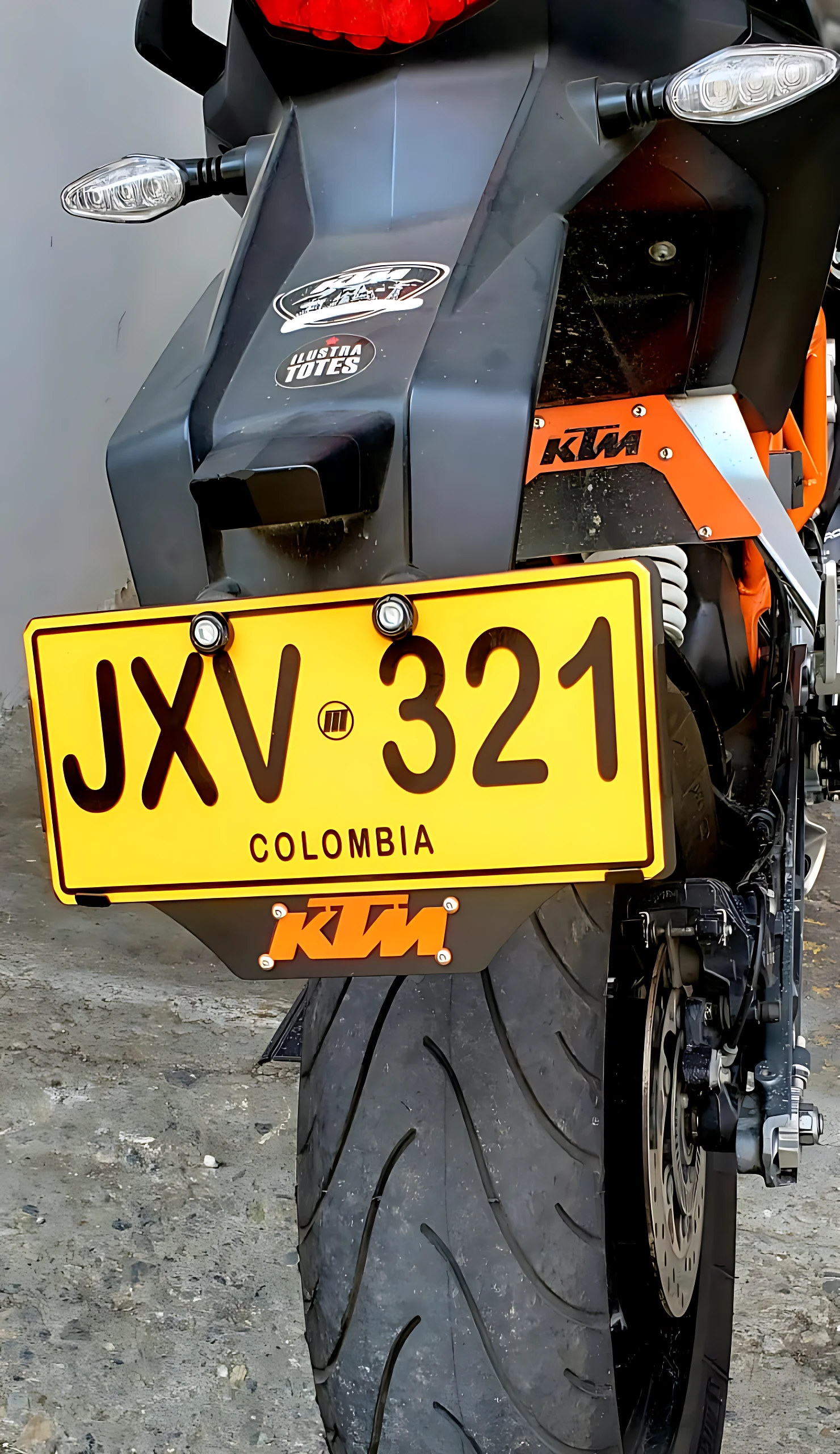 Portaplaca de acero inoxidable KTM 3D en negro mate, instalado en la parte trasera de una moto KTM naranja. Placa amarilla con texto negro JXV 321 y leyenda Colombia. Incluye luces LED laterales y logotipos oficiales.