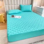 Protector de colchón acolchado en color turquesa con textura de rombos en relieve. Se muestra sobre una cama con dos almohadas del mismo diseño, acompañado de un libro y un reloj.