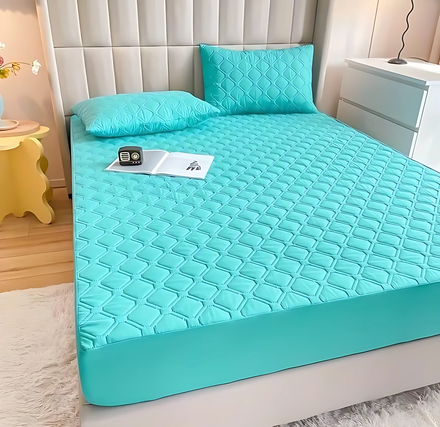 Protector de colchón acolchado en color turquesa con textura de rombos en relieve. Se muestra sobre una cama con dos almohadas del mismo diseño, acompañado de un libro y un reloj.