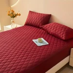 Protector de colchón rojo oscuro acolchado con costuras en rombos, sobre cama con dos almohadas a juego, con una taza sobre una revista en el centro.