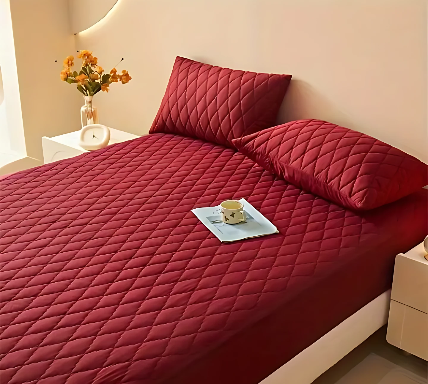 Protector de colchón rojo oscuro acolchado con costuras en rombos, sobre cama con dos almohadas a juego, con una taza sobre una revista en el centro.