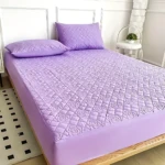 Protector de colchón acolchado color lila con patrón de rombos y relieve floral. Vista cenital sobre cama con dos almohadas del mismo tono y textura suave.